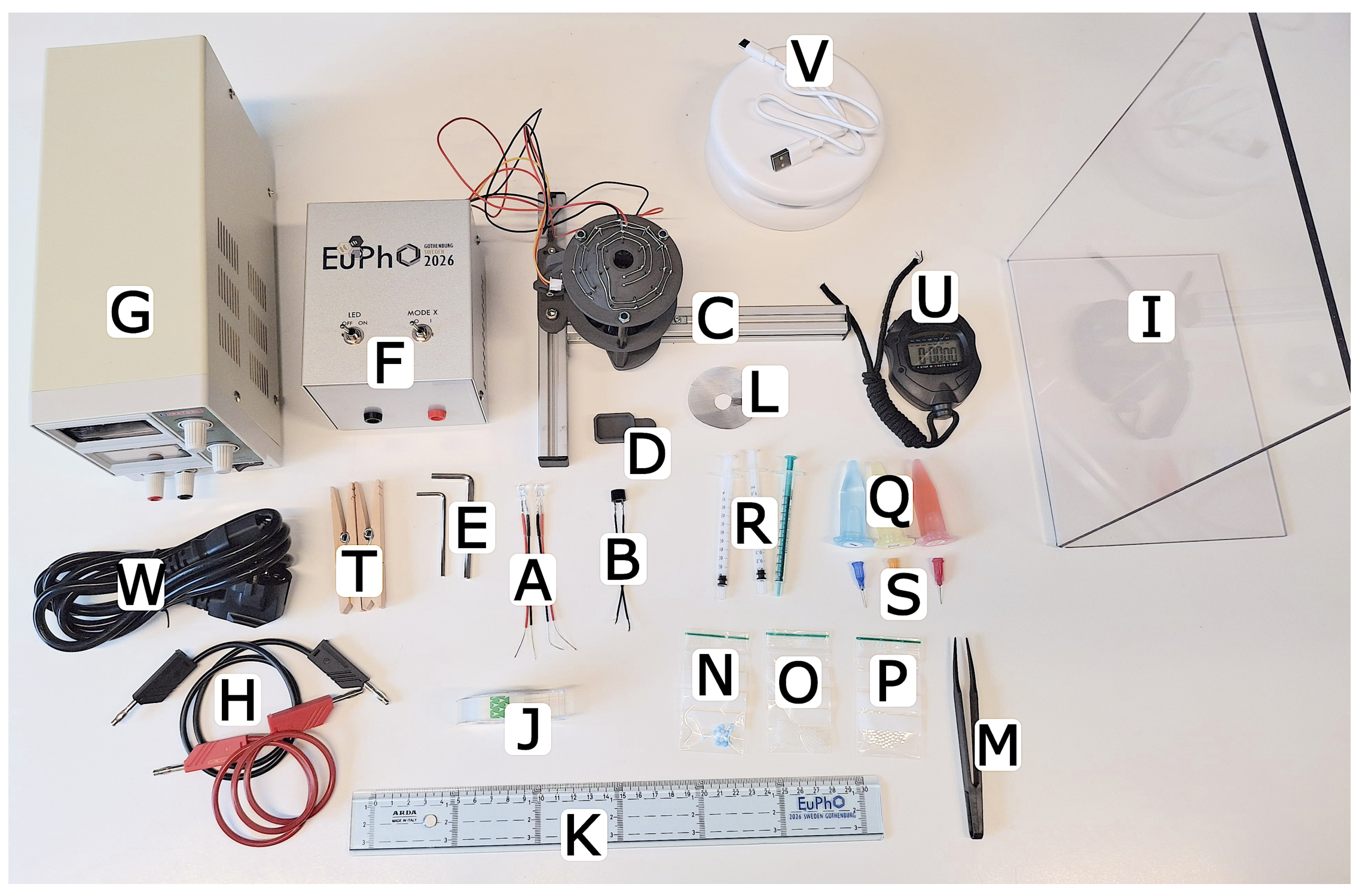 Photo of all the exam equipment laid out on a table, each item labelled with a white letter A through X: power supply, electrical box, levitator module, stopwatch, screen, syringes, liquids, tweezers, ruler, cables.