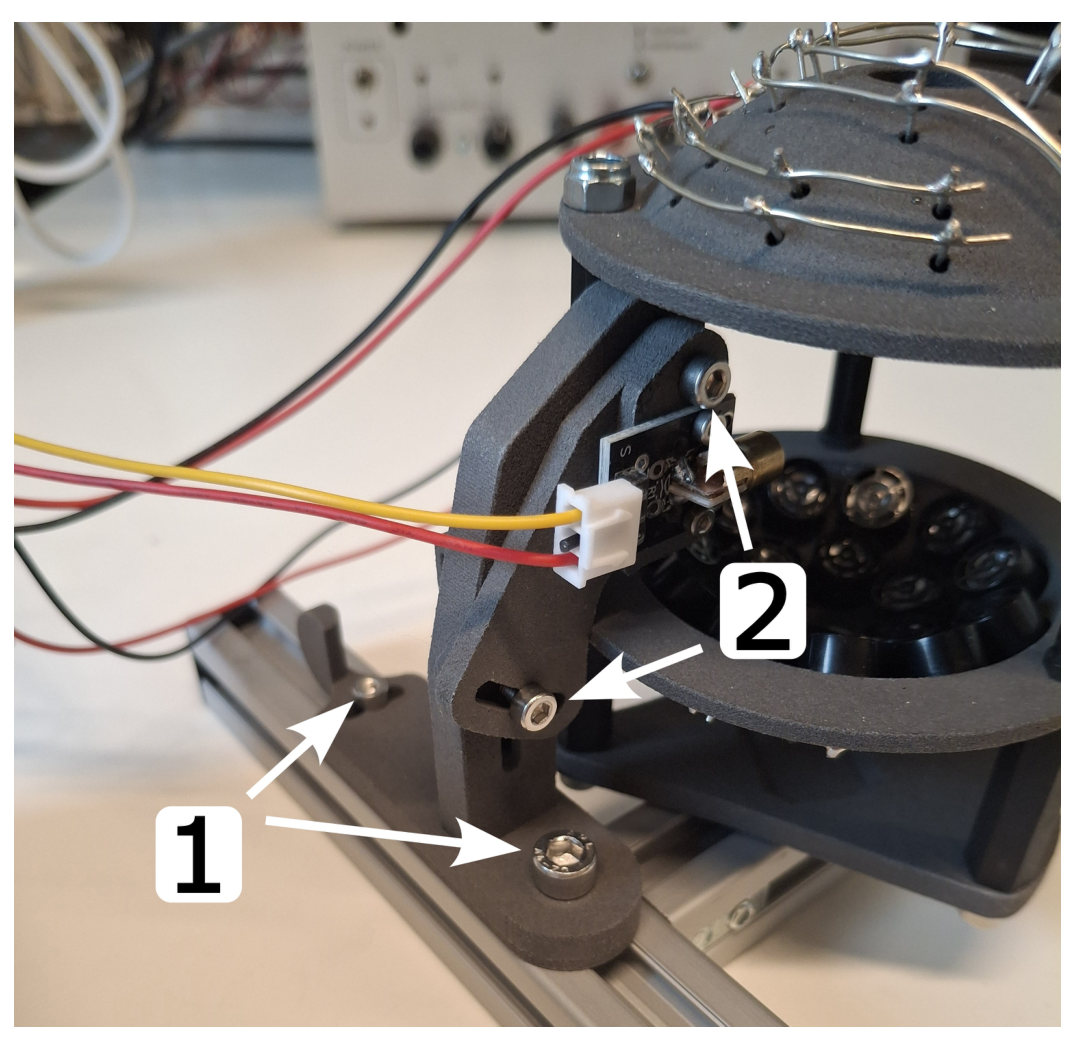 Photo of the levitator mounted on a rail with an LED module attached; white arrows labelled 1 point at two screws for horizontal movement and tilting, and arrows labelled 2 point at two screws for vertical movement and tilting of the LED module.