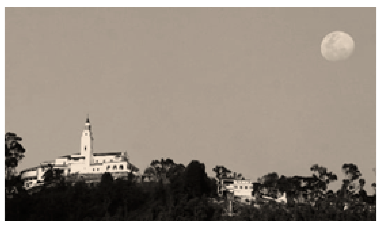 Photograph of the Moon low above the eastern hills at dusk, with the white sanctuary buildings of Monserrate on the hilltop.