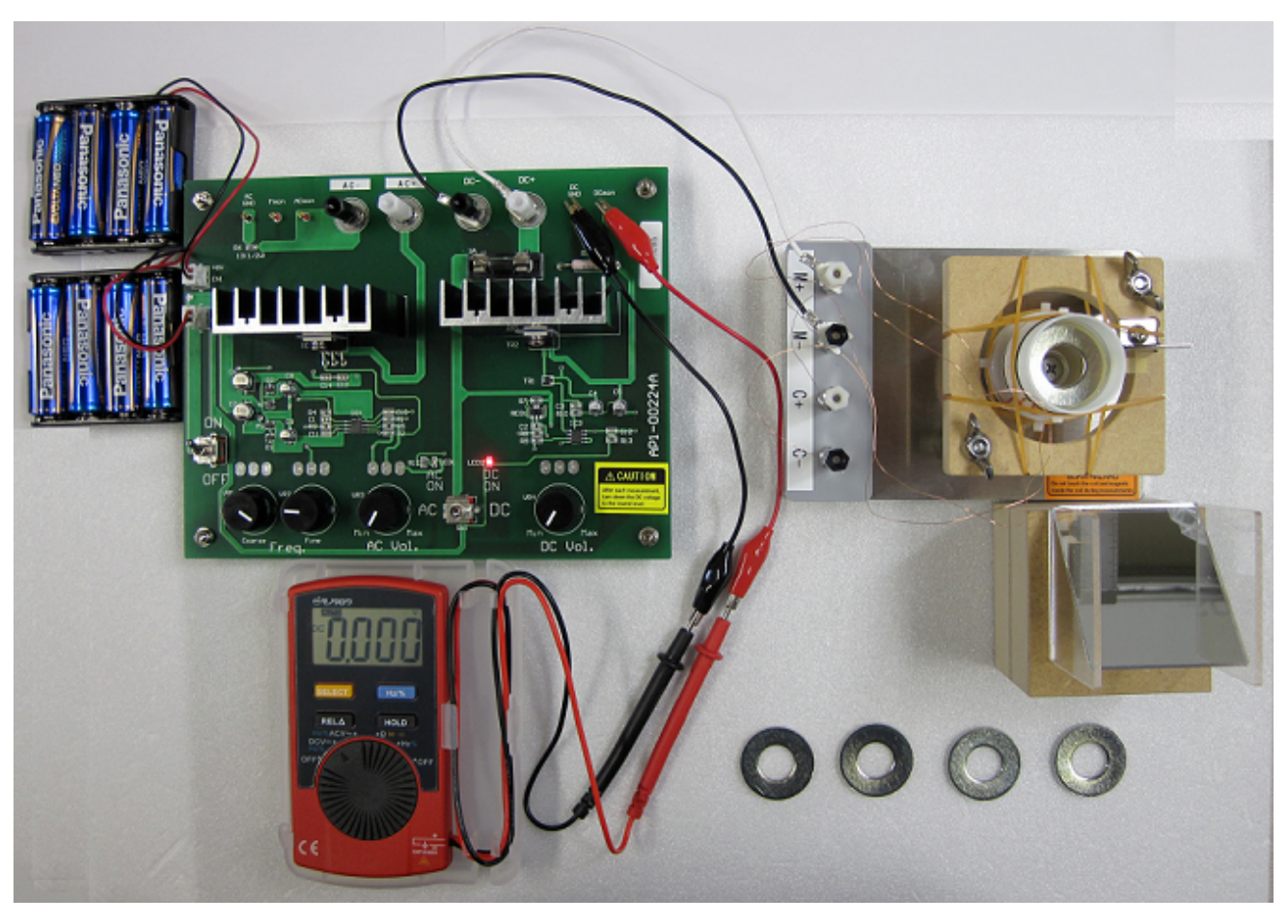 Photo of the experimental setup: power supply board with batteries, a DMM reading 0.000, alligator clip leads, the coil apparatus, a mirror, and four ring-shaped weights in front.