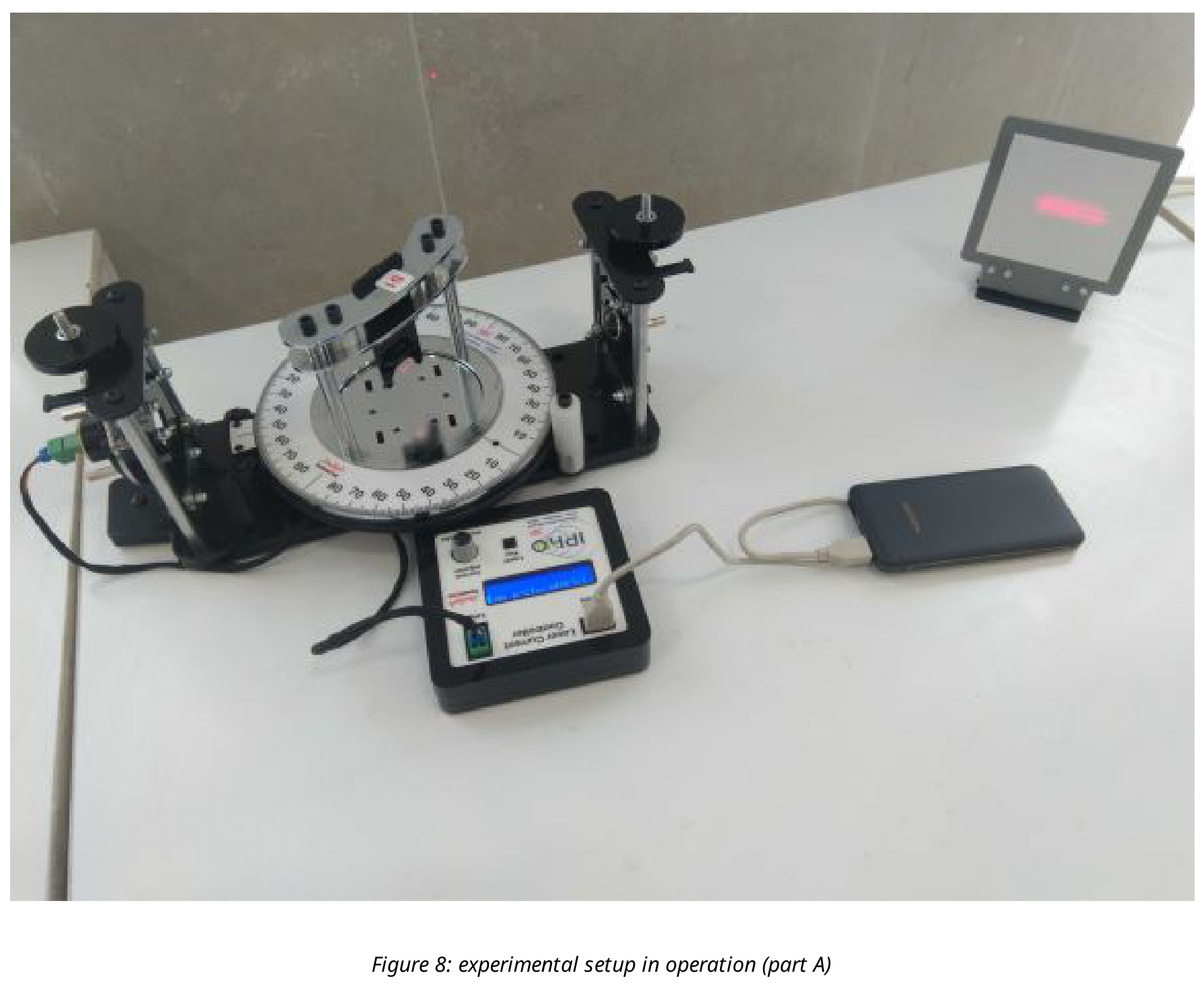 Photograph of the experimental table: a rotation protractor stage with a slide holder between a laser mount (left) and a screen with a red diffraction pattern (upper right); a laser power supply box with a display and a smartphone are on the table in front.
